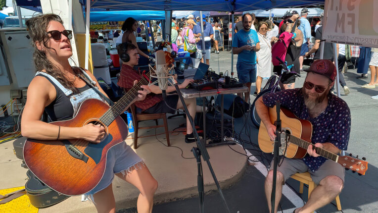 Outdoor performance with one musician standing and the other seated as others look on