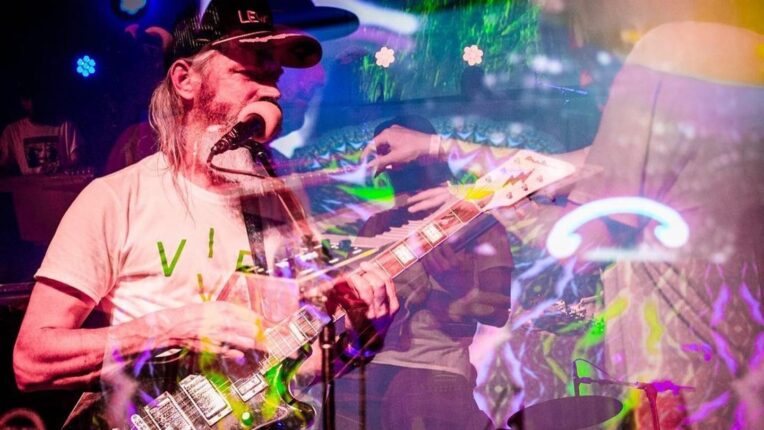 musician wearing baseball hat plays guitar with swirling light and the vague outlines of other musicians behind him.