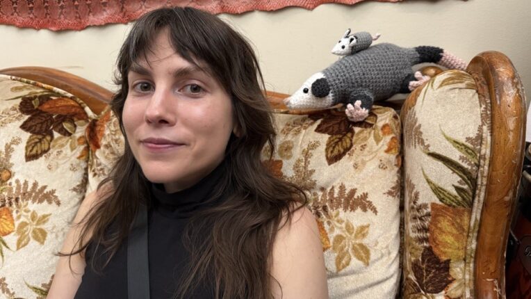 Woman with long brown hair sits on a couch with a crochet mother and baby opossum looking over her shoulder.