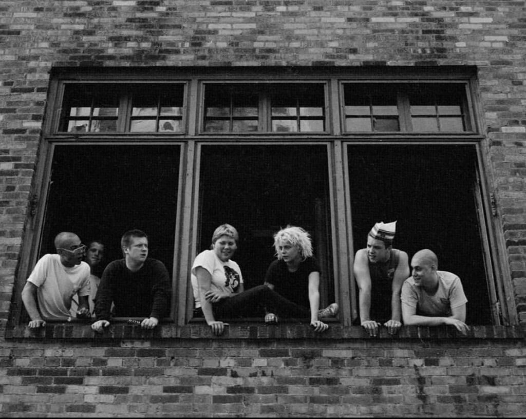 Seven young people lean and look out of three windows placed next to eachother in a brick building.