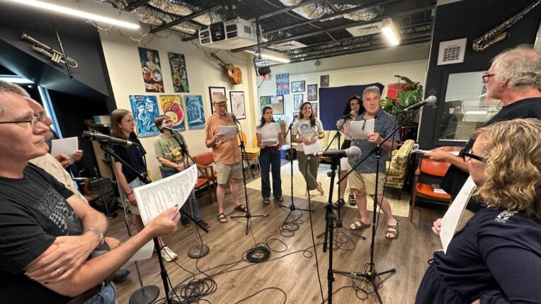 A group of men and women stand in a semi circle in front of microphones recording radio promos