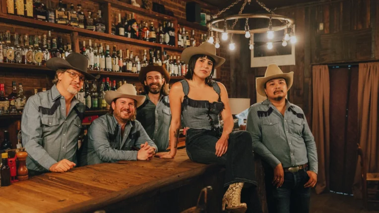 Woman with cowboy hat seated on bar with four men surrounding her