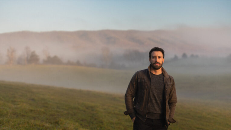 Bearded man standing in meadow with fog and mountain behind