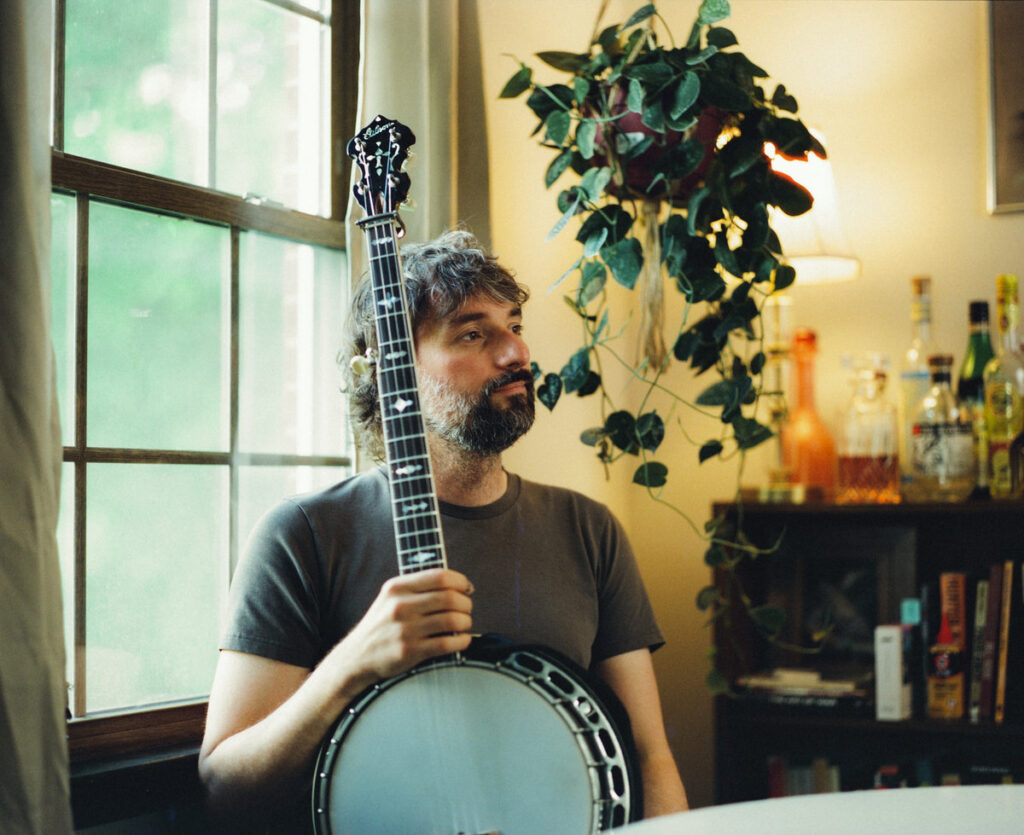 Man seated with banjo on knee in front of window and plant hanging on wall
