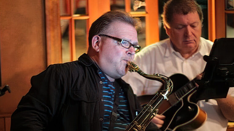 Man blowing saxophone with man playing guitar behind him