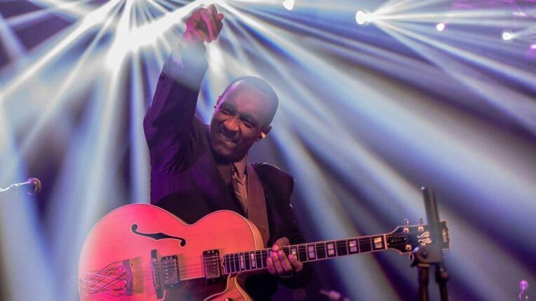 Man with arm raised holding guitar with light strobes around him