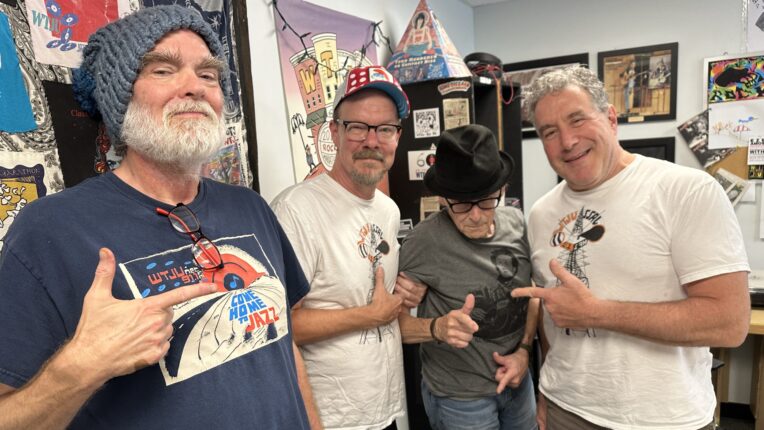 Four male DJs stand in a broadcast studio pointing at each other