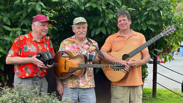Three men holding instruments standing in front of and behind bushes