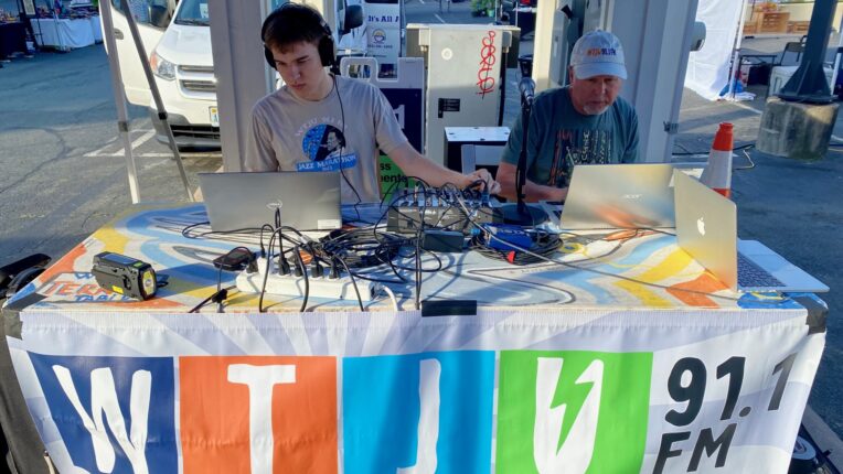 WTJU sound engineer Ben Larsen works a soundboard and laptop at the WTJU table at Charlottesville City Market. DJ Steve Kindig sits at a laptop next to him.