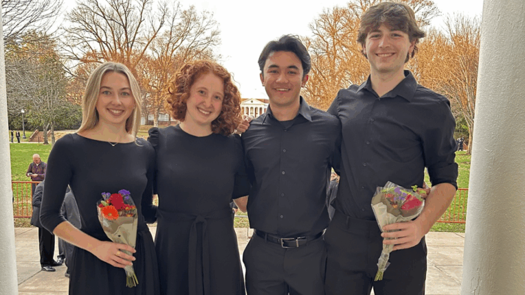 Four students in black smile for the camera in front of autumnal trees. Two students hold a bouquet of flowers.