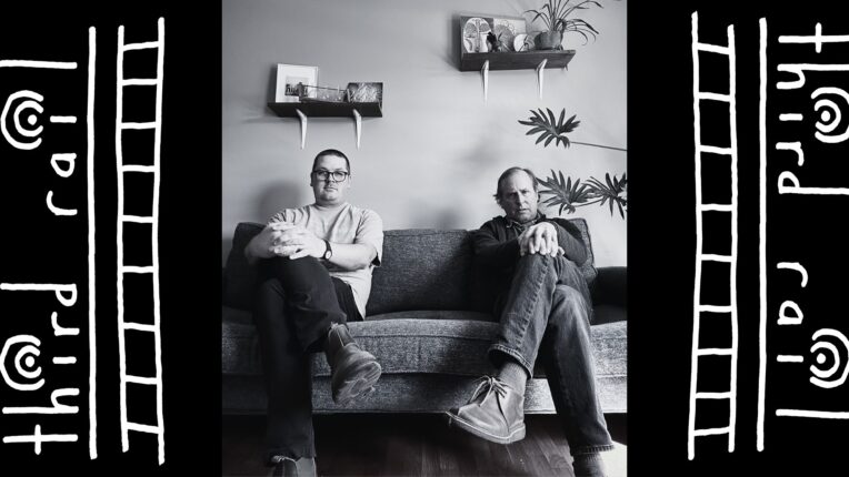Jordan Perry and Ned Oldham sit on a couch with their legs crossed, looking at the camera.
