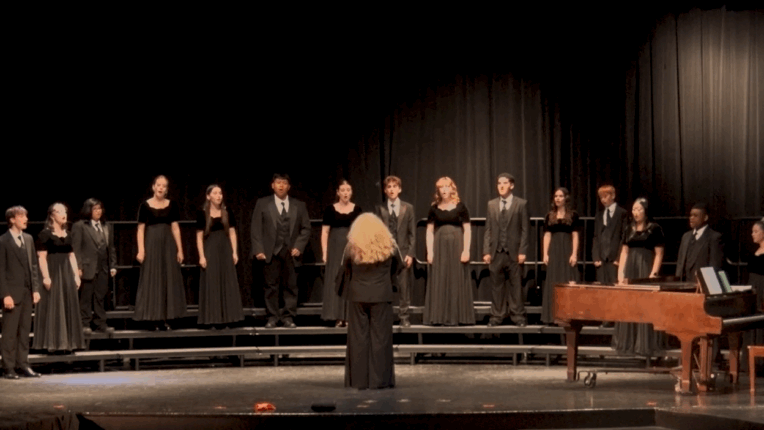 A choir in black clothing stands on a stage in a semi-circle, with a director standing in front of them at the center. There is a piano and pianist on the right side of the stage.