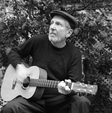 Man with cap seated on chair outside while holding a guitar