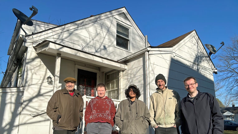 Five people standing in front of a white house with a clear blue sky above