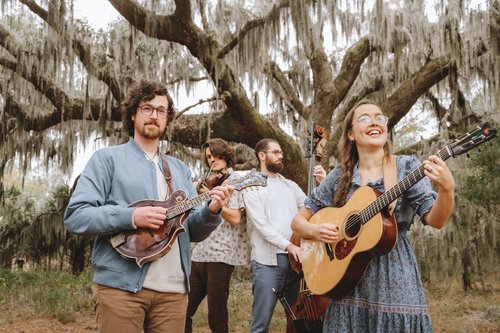 Four musicians holding instruments outside in front of a tree with moss hanging over it