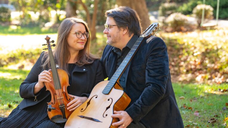 Woman holding violin and man holding guitar with both seated outside on a sunny day in front of a tree