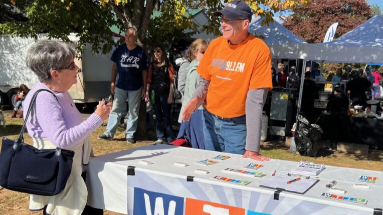Daniel Hennicke in an orange shirt and baseball cap stands at the WTJU table speaking to a woman who has stopped by