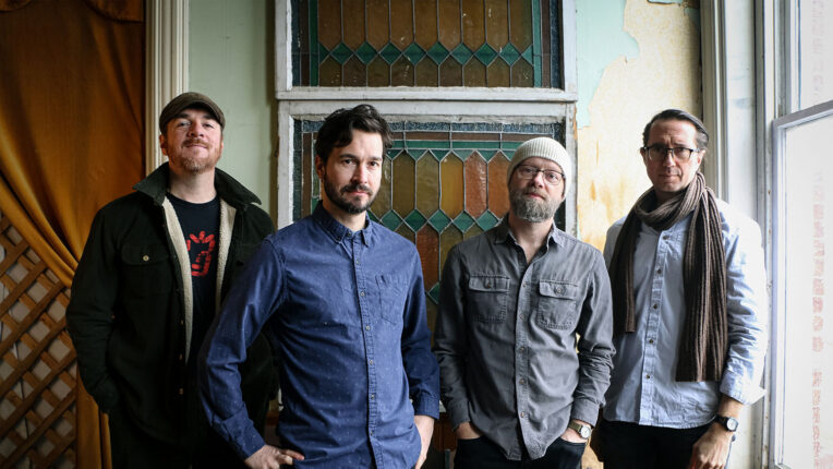 Four men standing inside with door behind and a window to the side