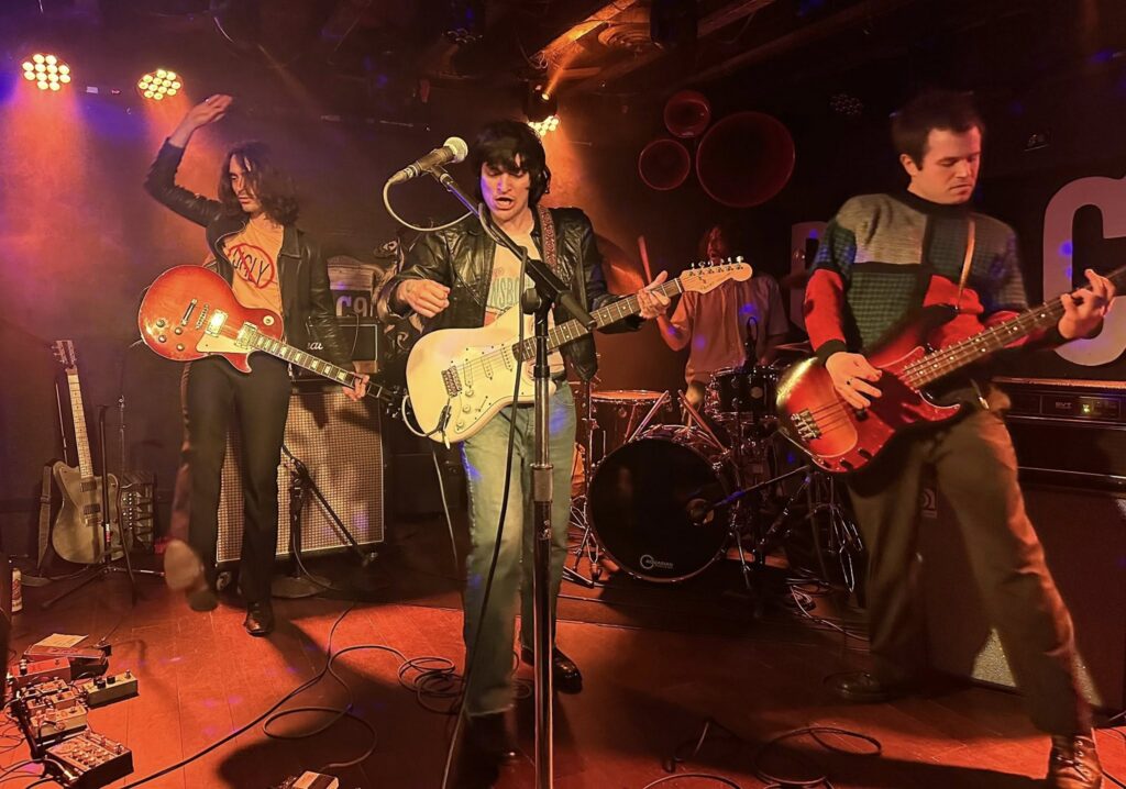 Rock band Native Sun playis music on a stage, two guitar players and a bass player strumming their instruments, with a drummer in the background, all under stage lighting.