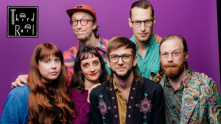 Six members of the North Country band stand in front of a purple wall in colorful clothes. A logo for Third Rail is in the left hand corner.