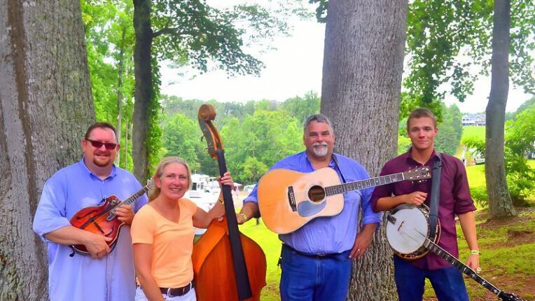 four-piece musical group posed outdoors, holding string instruments typical of the bluegrass genre