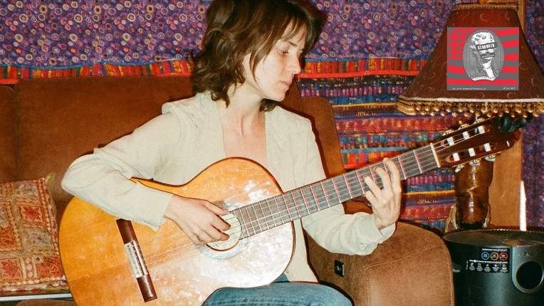 Musician Jana Horn sits on a brown couch playing an acoustic guitar with a colorful wall behind her.