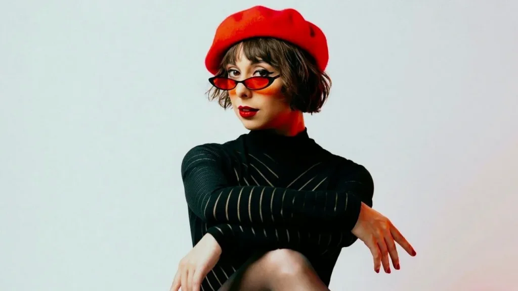 Musician Veronica Swift sits with legs and arms crossed, wearing a red hat and red sunglasses.