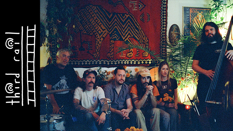 Five members of band mead the dear sit on a couch with another member standing up with a stand up bass. There is a tapestry and some plants behind them, and the decorative logo and words 