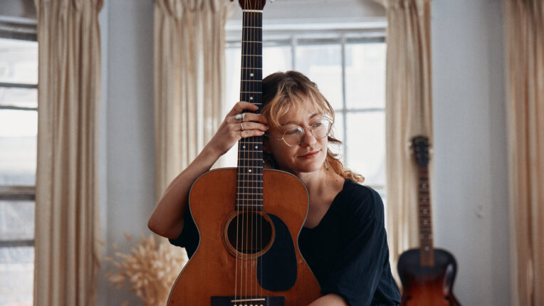 Female musicians seated holding guitar upright with another guitar in the background