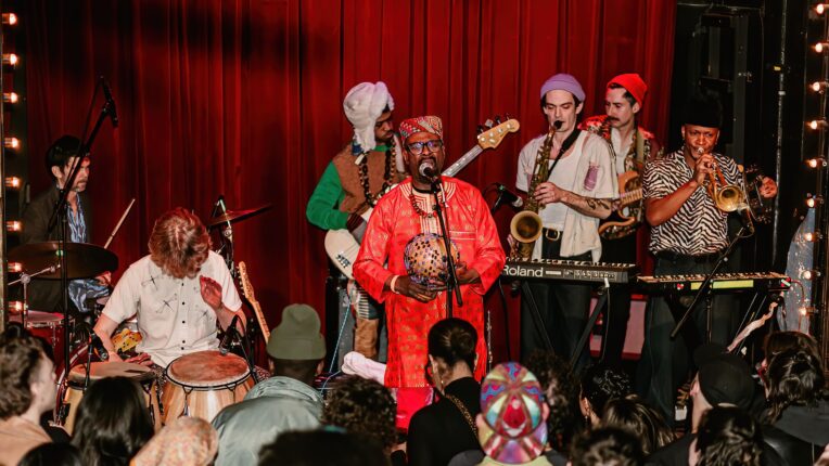 Super Yamba Band performing live, featuring a brightly colored red dashiki and intricate jewelry