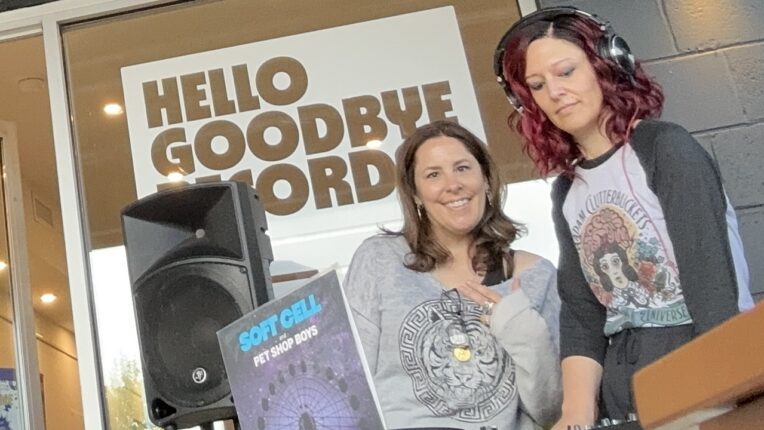 Two DJs spin records in front of a sign that says Hello Goodbye Records.