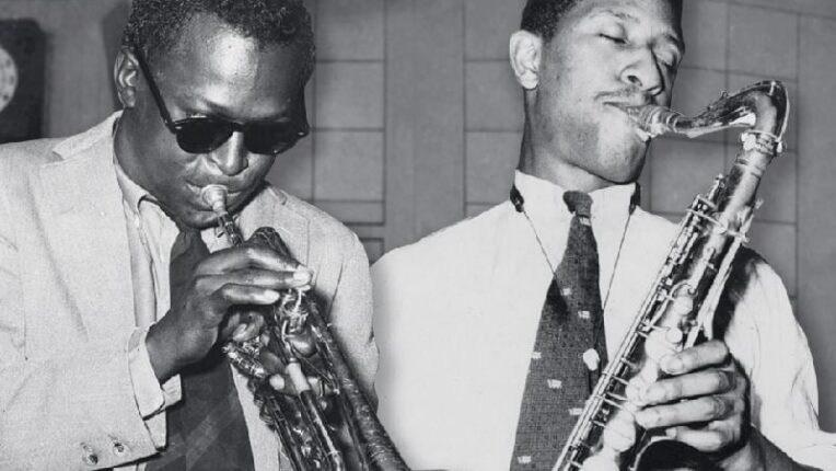 Miles Davis and Sonny Rollins stand side by side, blowing their horns