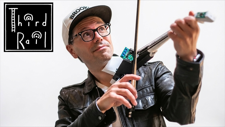 Musician Brian Lindgren, in a baseball cap, plays an electronic viola. The log for Third Rail is in the top left corner.