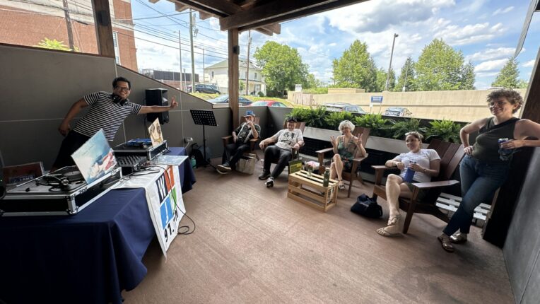 A vinyl DJ flashes the peace sign. Four people sit on chairs in front of him, and one person stands, on a balcony with blue skies, scattered clouds, and trees in the background.