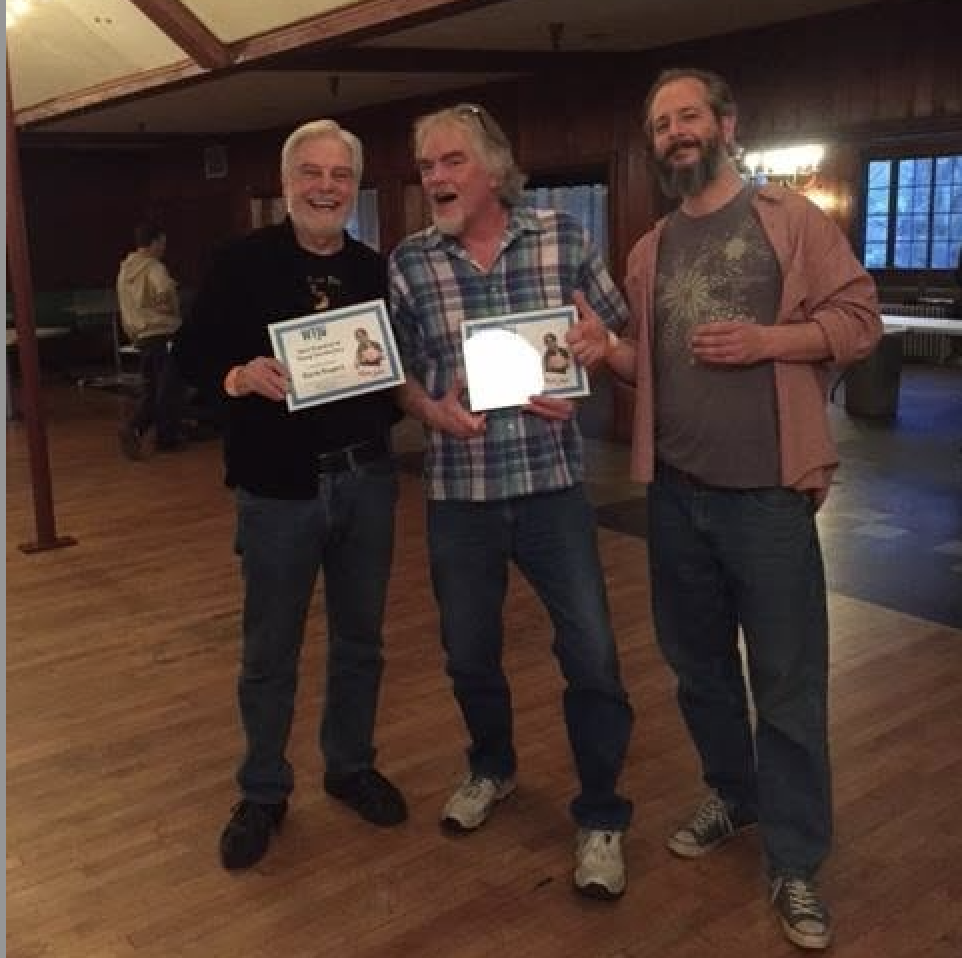 Three men stand next to eachother in a dimly lit room. Two of them hold awards.