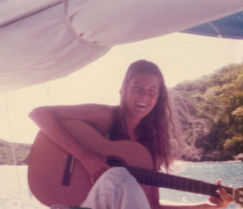 Musician Cathy Hamer sits on a boat smiling and holding an acoustic guitar. Water and hills are in the backdrop.