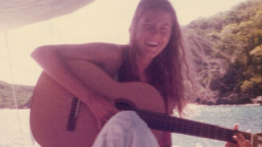Musician Cathy Hamer sits on a boat smiling and holding an acoustic guitar. Water and hills are in the backdrop.
