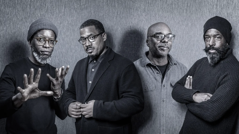 Black and white photo of four men in varying positions standing against a gray wall.