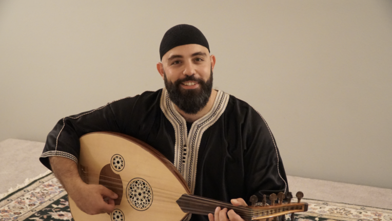 Abed Alsalam Manasra sits on an ornate rug, smiling and holding an oud