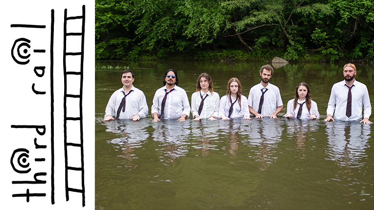 Seven people in white shirts and black ties stand in murky water with trees behind them.
