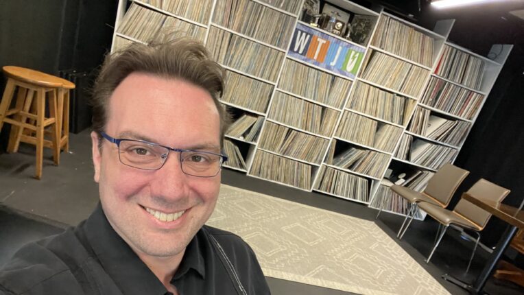 General manager of WTJU, Nathan Moore, stands in front of the WTJU stage and a wall of vinyl records.