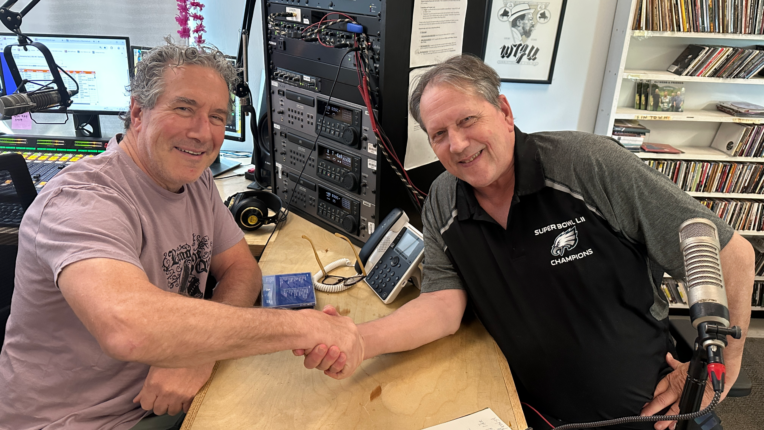 Radio hosts Brian Keena and Charlie Curtis grasp hands over across a tble and smile for the camera