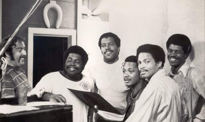 six men stand around a music stand, mostly smiling at the camera