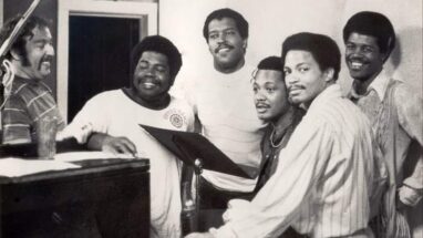six men stand around a music stand, mostly smiling at the camera