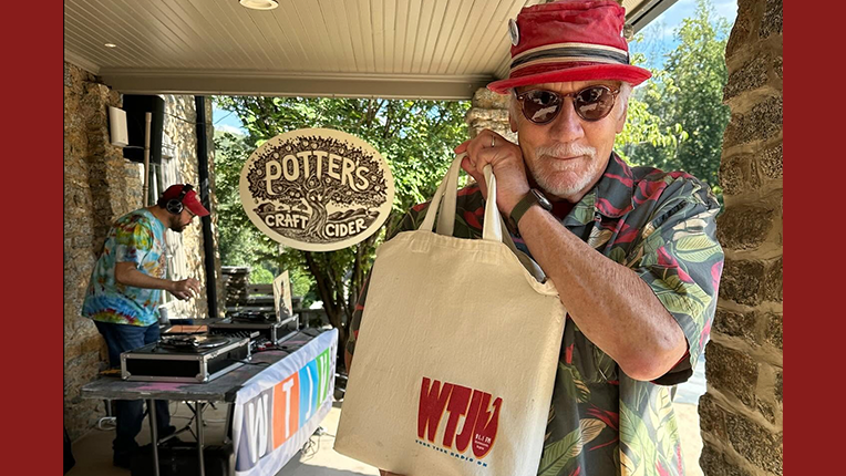 DJ in red hat stands holding a white tote bag that says WTJU. Behind him a DJ spins vinyl records.
