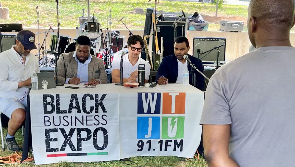 Photo of the Business Pitch Competition at the 2026 Black Business Expo features four judges writing down notes while a man presents his business idea.