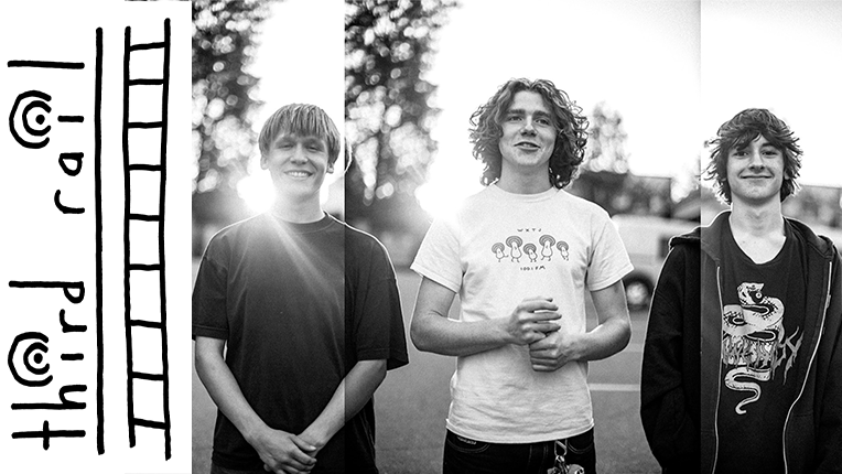 Three young men stand in line smiling at the camera. The sun shines through trees behind them.