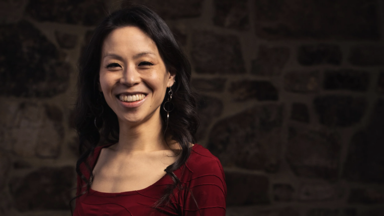 Pianist, Jeannette Fang, in a red top, stands in front of a stone wall, smiling at the camera.