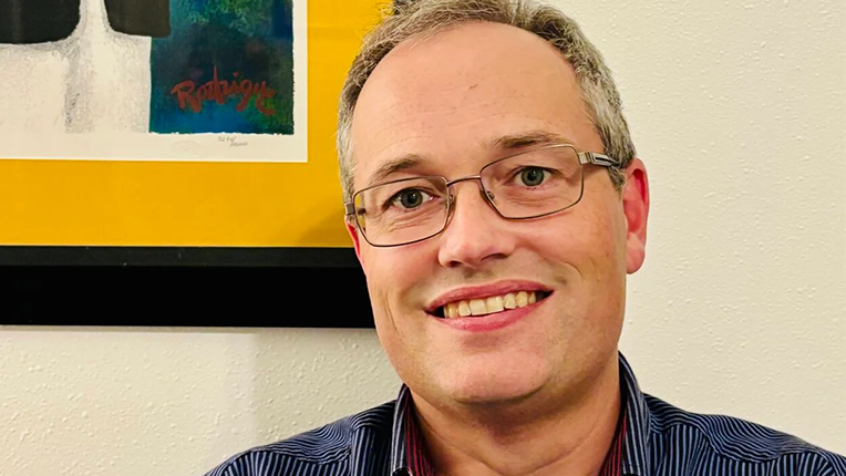 Man smiles at the camera in front of the corner of a bright framed photo and a white wall.