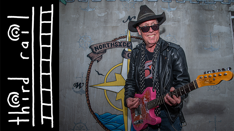 Guitarist Jimmy Catlett holds a pink guitar and smiles at the camera. He is wearthing a leather jacket and a hat.